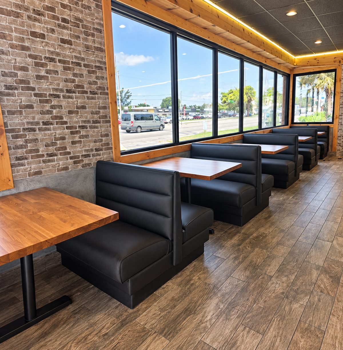 Row of black upholstered restaurant booths with wood tables beside a wall of windows.
